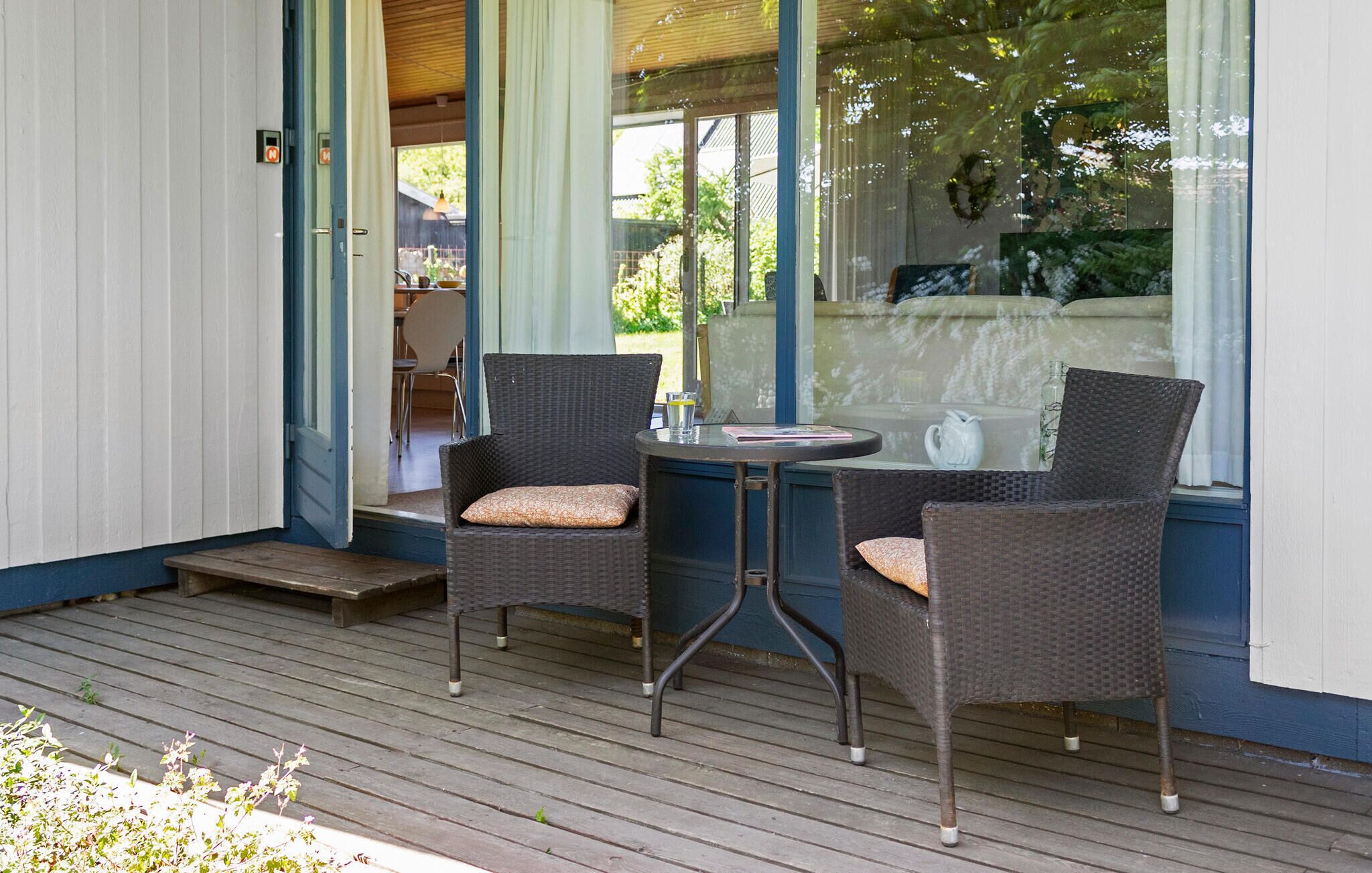 A covered outdoor seating area with wicker chairs and a small table, a wooden deck, and a view of the outdoors through a glass door.