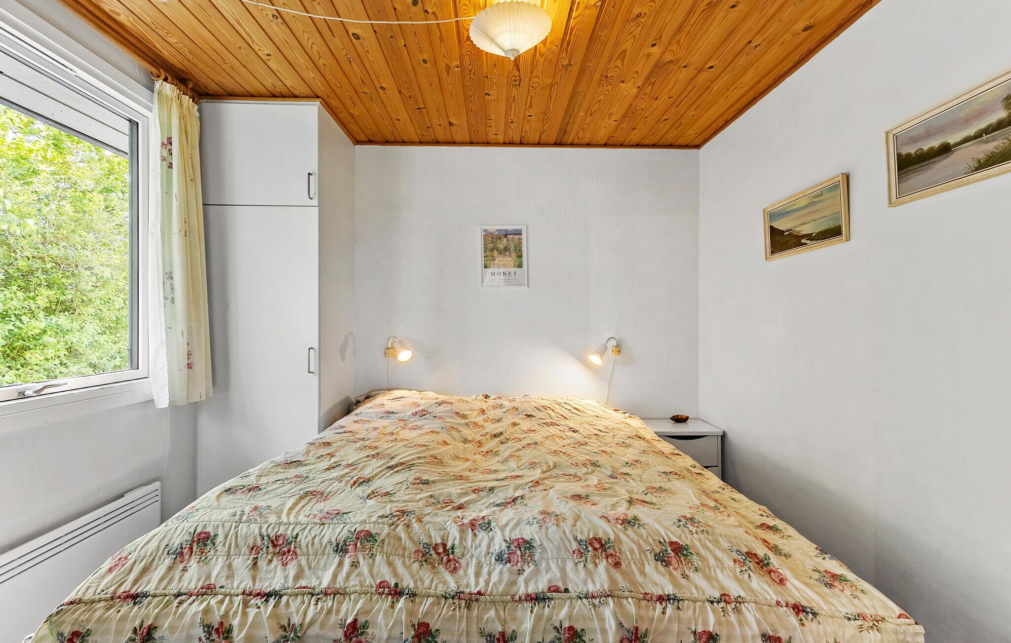 Un dormitorio con techo de madera, una cama con colcha estampada, dos lámparas de pared, un cuadro enmarcado y una ventana con cortinas.
