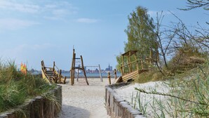 A sandy beach path leading to a wooden structure with a swing set, surrounded by grass and trees, overlooking a body of water with buildings in the distance. - Ferienhaus 6 Altefähr (Altefähr/Rügen)