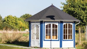 Un petit kiosque de plain-pied, doté d’un toit en pente et de grandes fenêtres, entouré de pelouse et d’arbres.