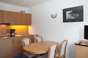 A compact kitchen with wooden cabinets, a round dining table, and chairs. There is a coffee maker, a sink, and a framed picture on the wall.