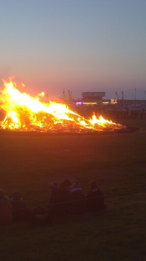 A large fire with flames and smoke, a building in the background, and a group of people sitting on the ground.