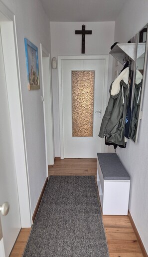 A hallway with a wooden floor, a grey rug, a white door with a textured glass panel, a black cross on the wall, and a coat rack with jackets. - North Sea House Dornumersiel - house (Dornum)