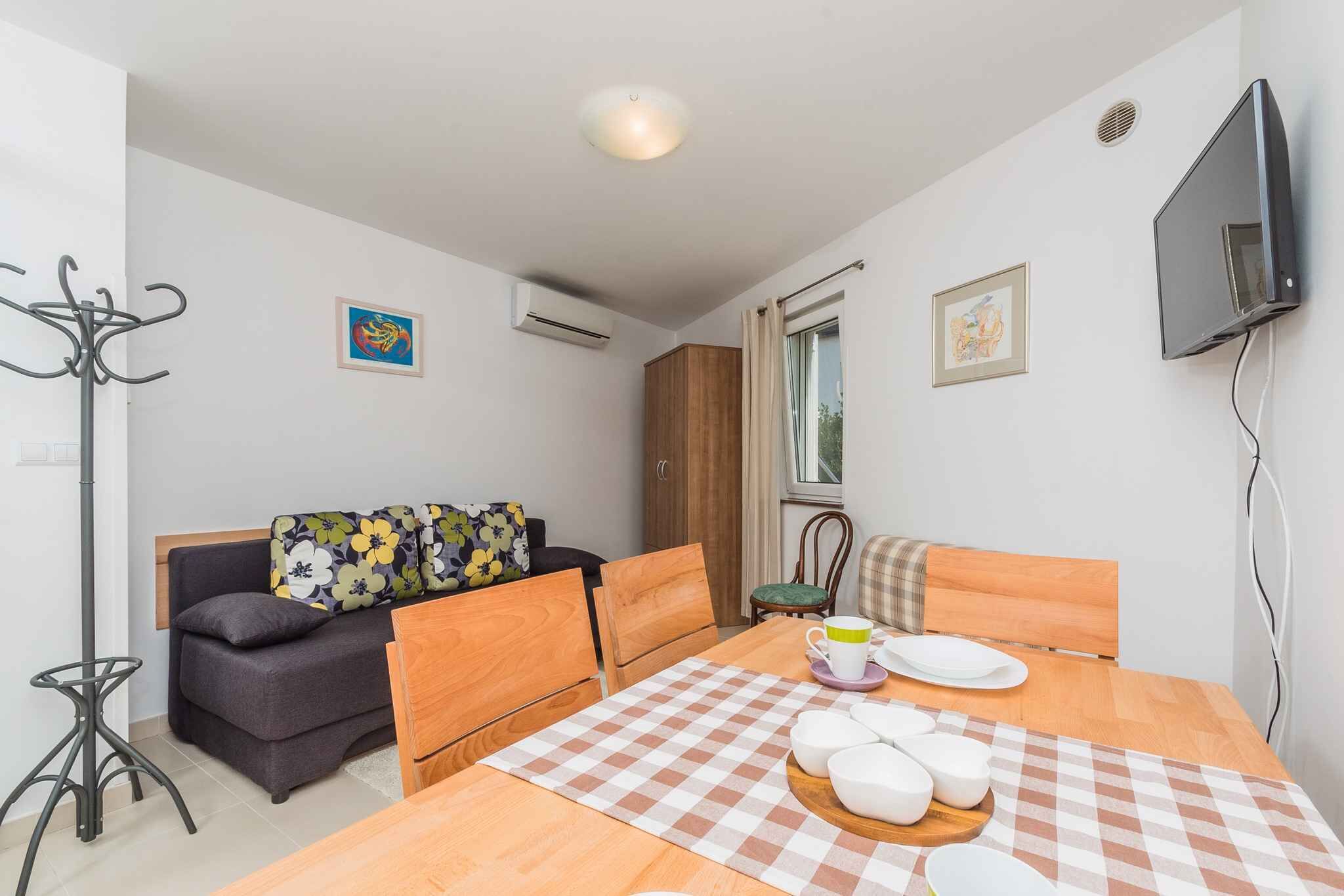 Un espacio habitable compacto con un sofá, una mesa de comedor con mantel a cuadros, un televisor montado en la pared y una ventana con cortinas.