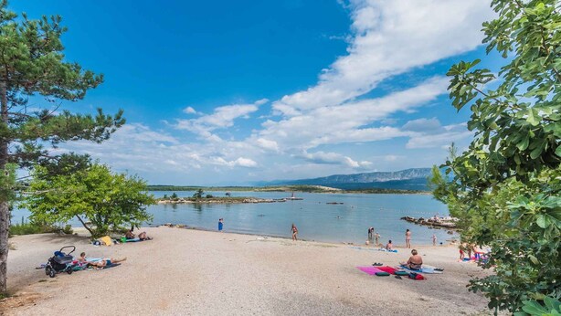 Ein Strand mit badenden und sonnenden Menschen, umgeben von Bäumen und Bergen im Hintergrund.