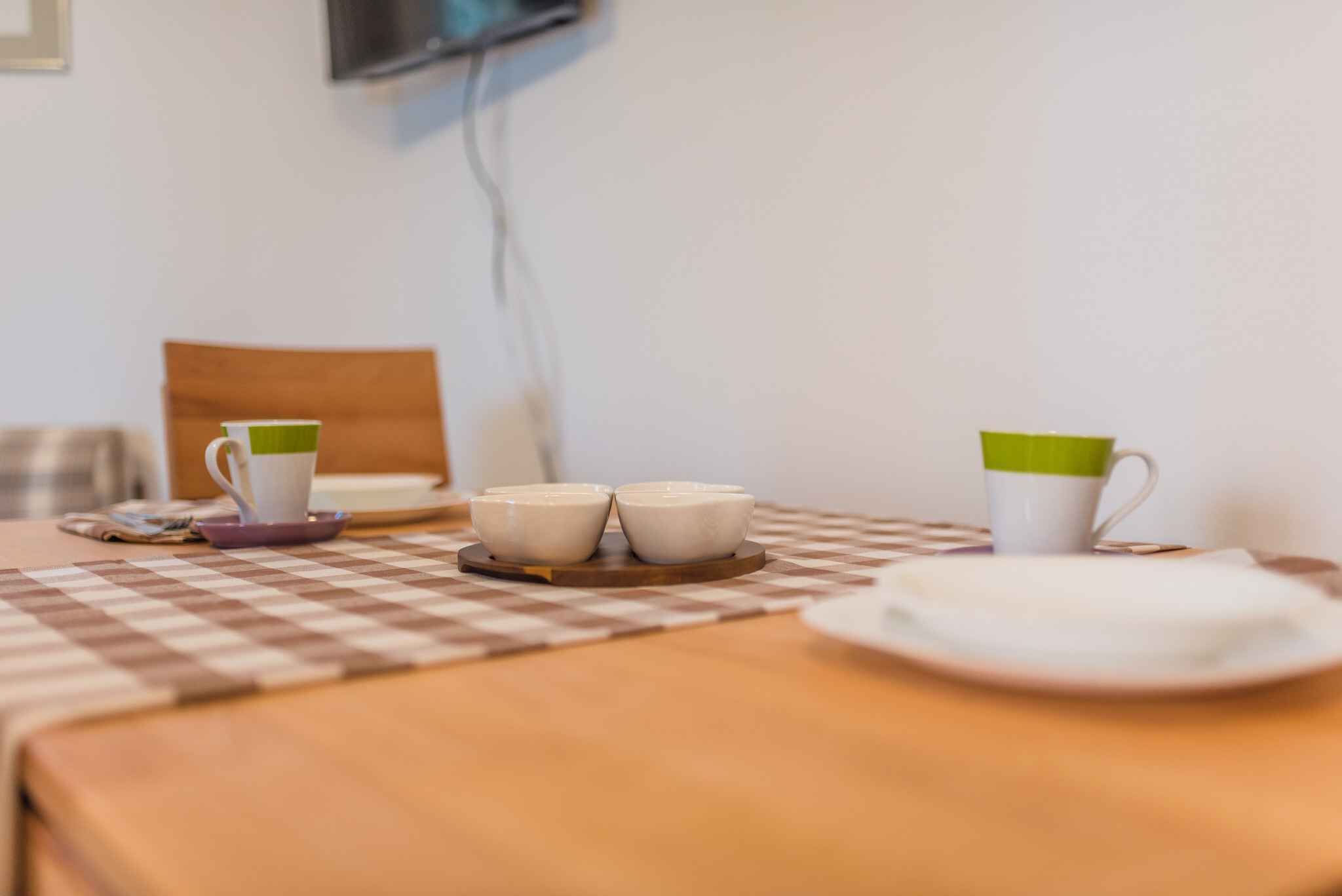 A checkered table with two white cups and saucers, a plate, and a television mounted on the wall.