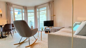 Ein modernes Wohnzimmer mit grauem Sofa, einem Schaukelstuhl, einem Flachbildfernseher und großen Fenstern mit Vorhängen.