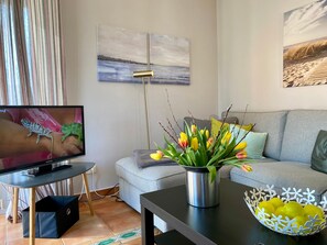 Ein Wohnzimmer mit Fernseher, einer Couch, einem Couchtisch mit Blumen und einer Obstschale.