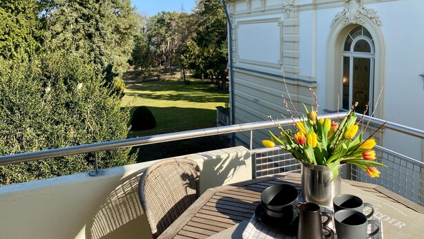 Ein Balkon mit Blick auf einen gepflegten Rasen und Bäume, ein Korbstuhl, ein Metalltisch mit Tablett und eine Vase mit Blumen.