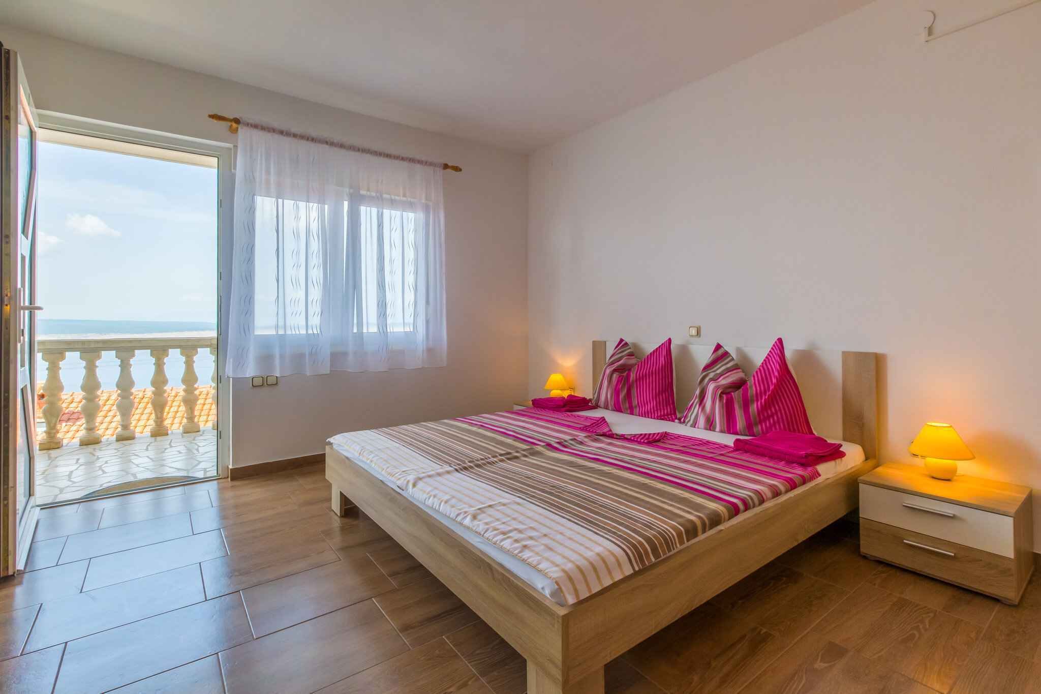 A bedroom with a wooden bed, a nightstand with a lamp, a window with curtains, and a balcony with a railing.