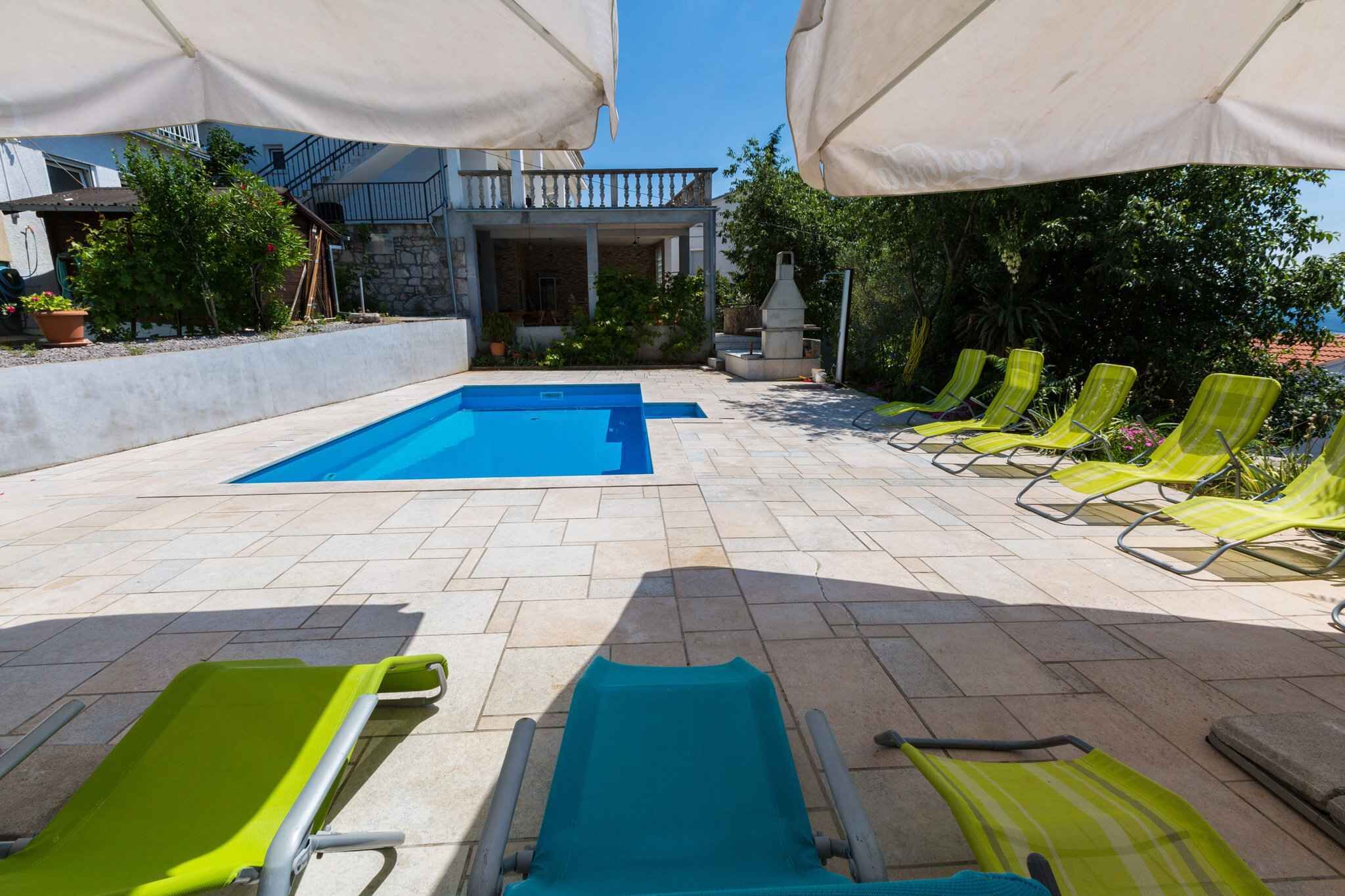A swimming pool with adjacent sun loungers and umbrellas, surrounded by a stone patio and greenery.