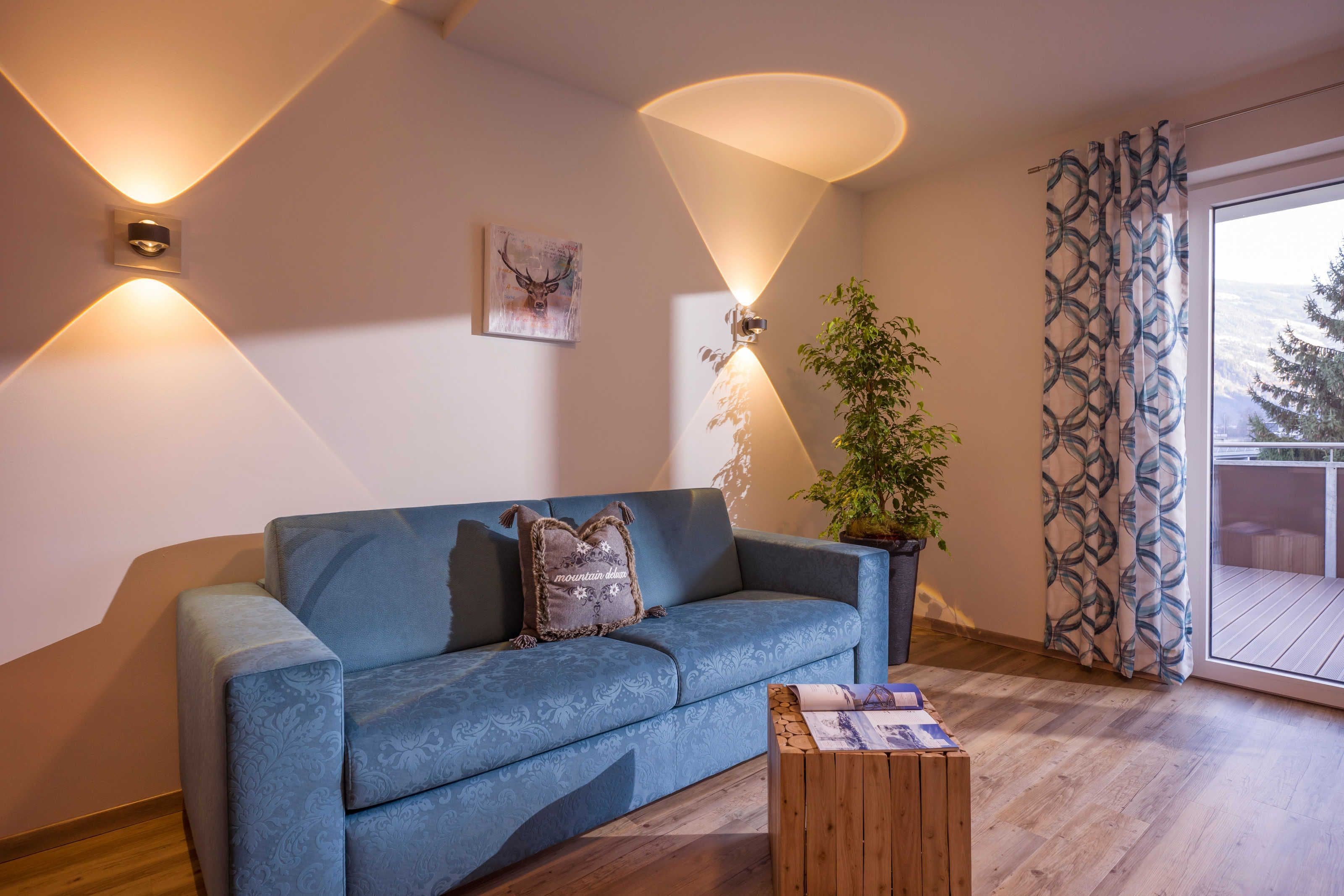 Un sofá azul con un cojín, una mesa de centro de madera, una planta en maceta y un aplique de pared.