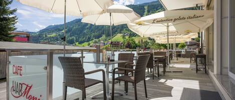 An outdoor cafe with wicker chairs and large umbrellas, overlooking a mountainous landscape.