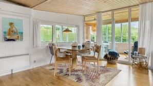 A spacious room with a wooden ceiling, large windows, a dining area with chairs and a table, and a view of greenery outside.