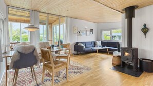 A spacious living room with a large wooden ceiling, a sofa, a dining table with chairs, and a wood-burning stove.