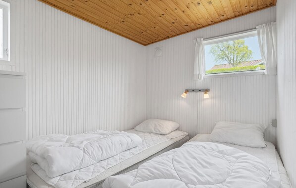 A room with two single beds, a wooden ceiling, a window with curtains, and a wall-mounted light fixture.