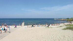 A beach with a wooden walkway leading to the water, people sunbathing, and boats in the background.