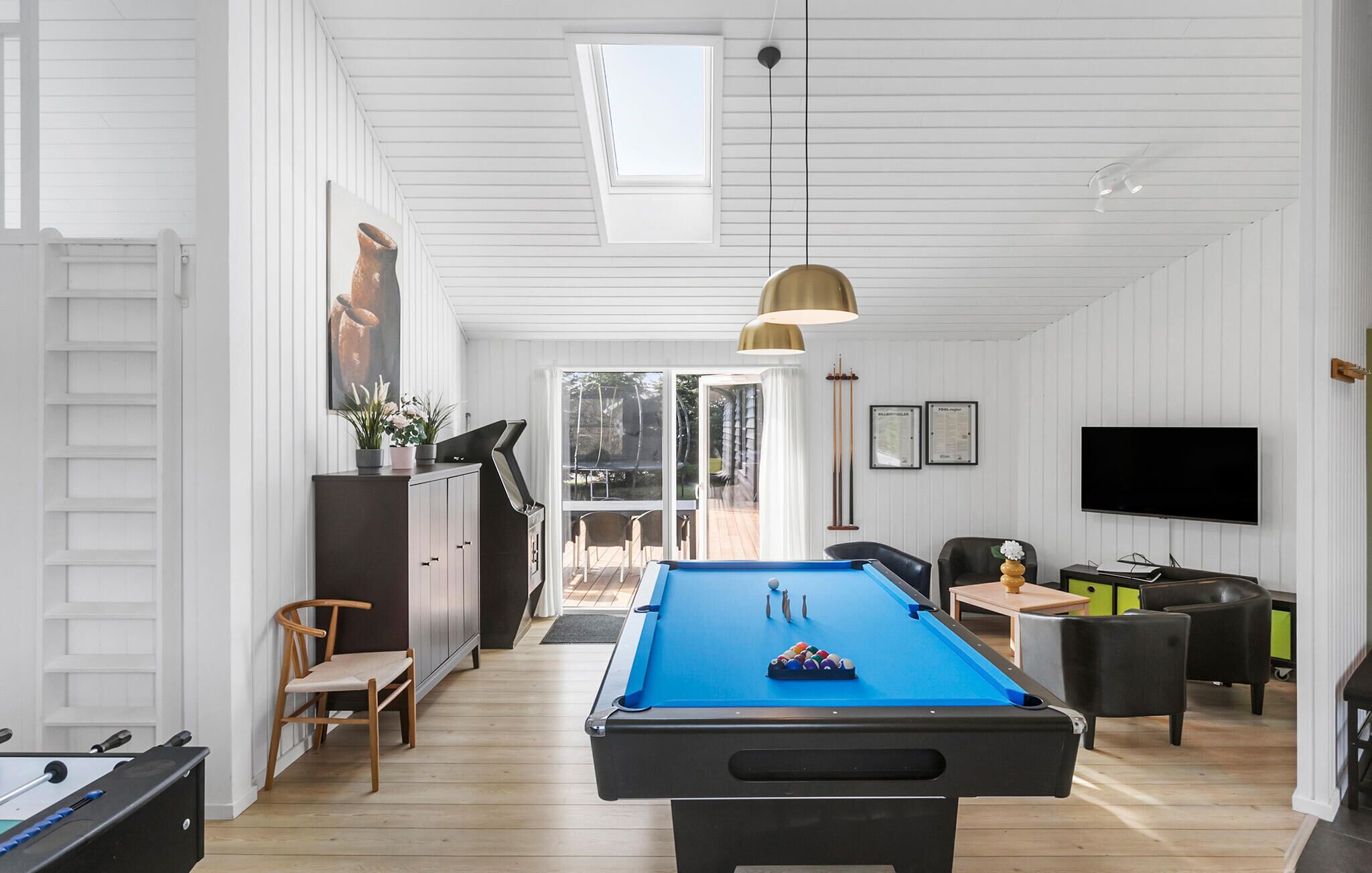 Une table de billard dans une pièce aux murs blancs et au plancher en bois, avec un téléviseur et un coin repas comprenant des chaises et une table.