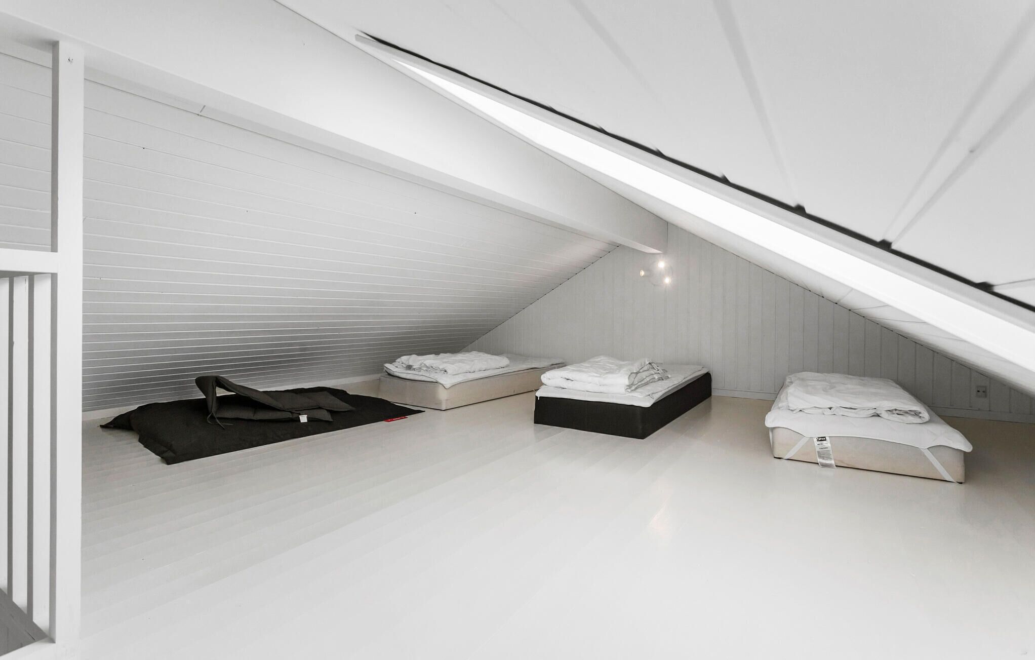 A small, white attic bedroom with three beds arranged in a row, a black chair, and a white ceiling with a skylight.