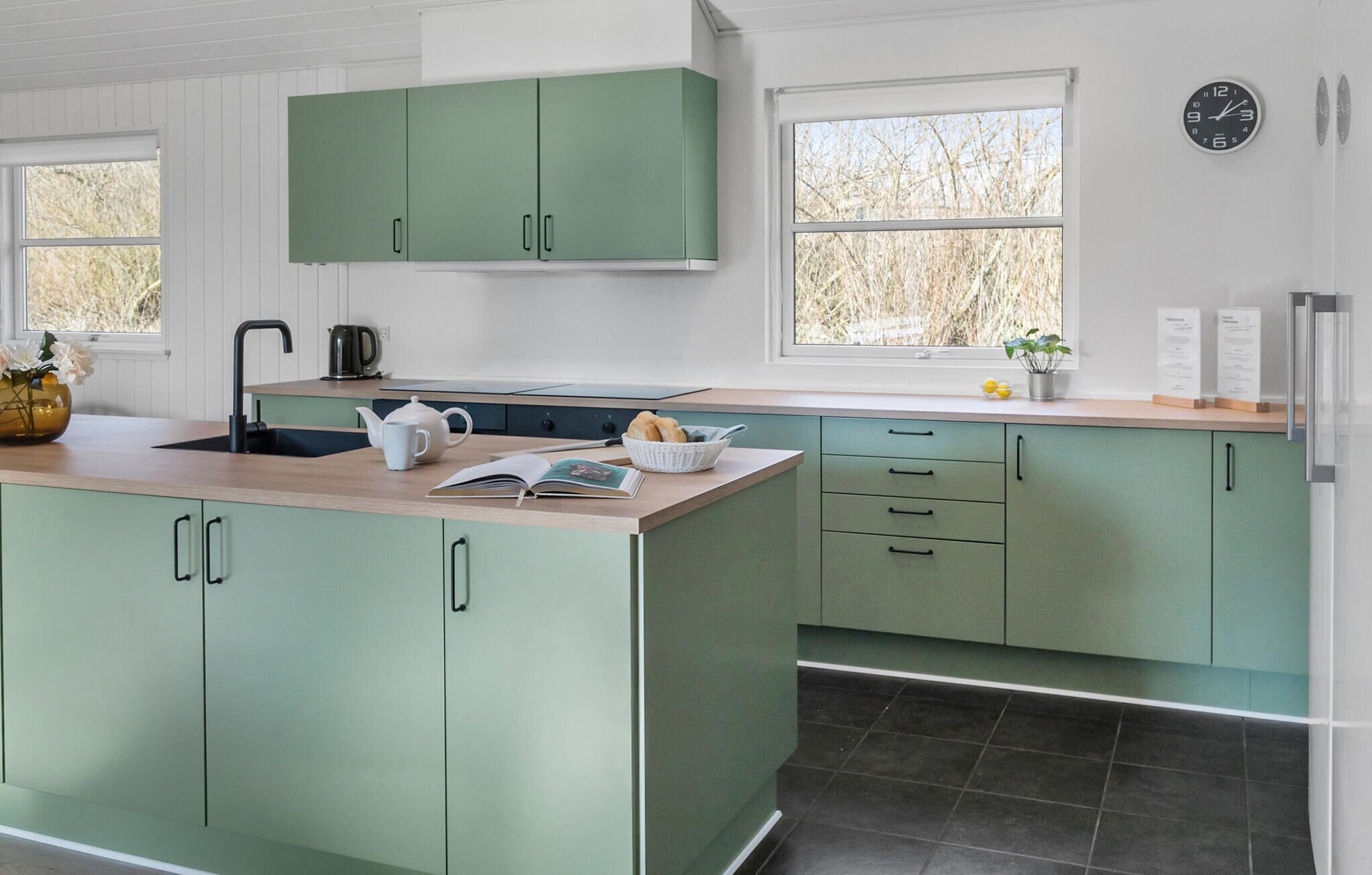 A kitchen with green cabinets, a black sink, a kettle, a basket of bread, and a clock on the wall.