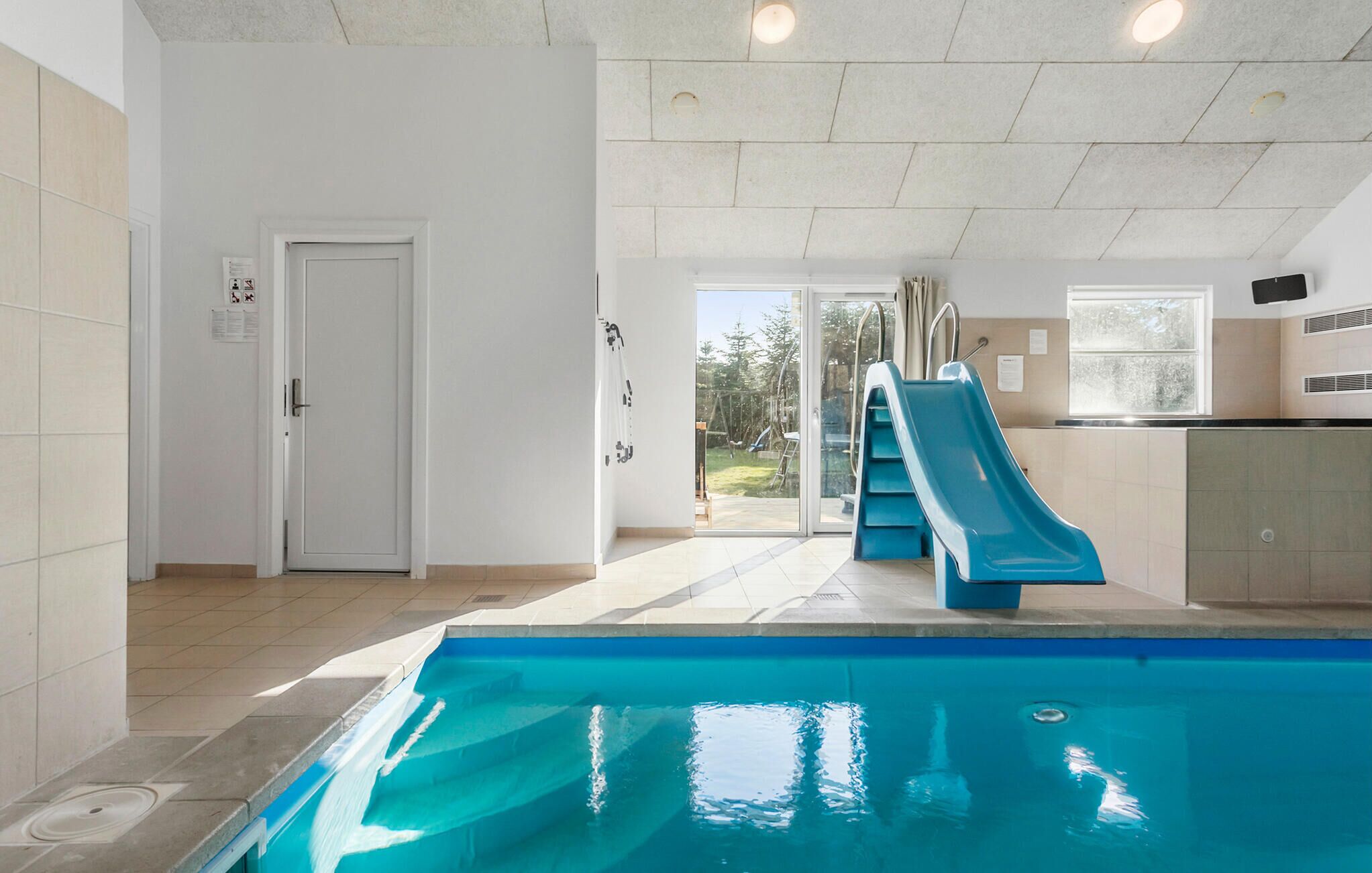 A swimming pool with a blue slide, a door, and a view of the outside.