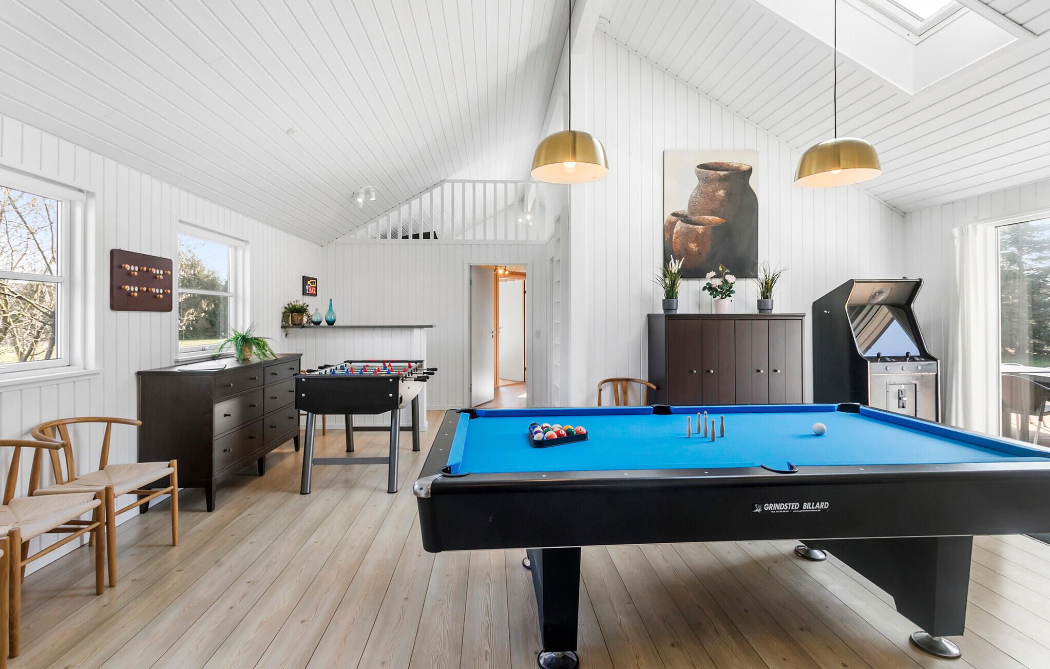 A billiards room with a pool table, two hanging pendant lights, a foosball table, and a grill.