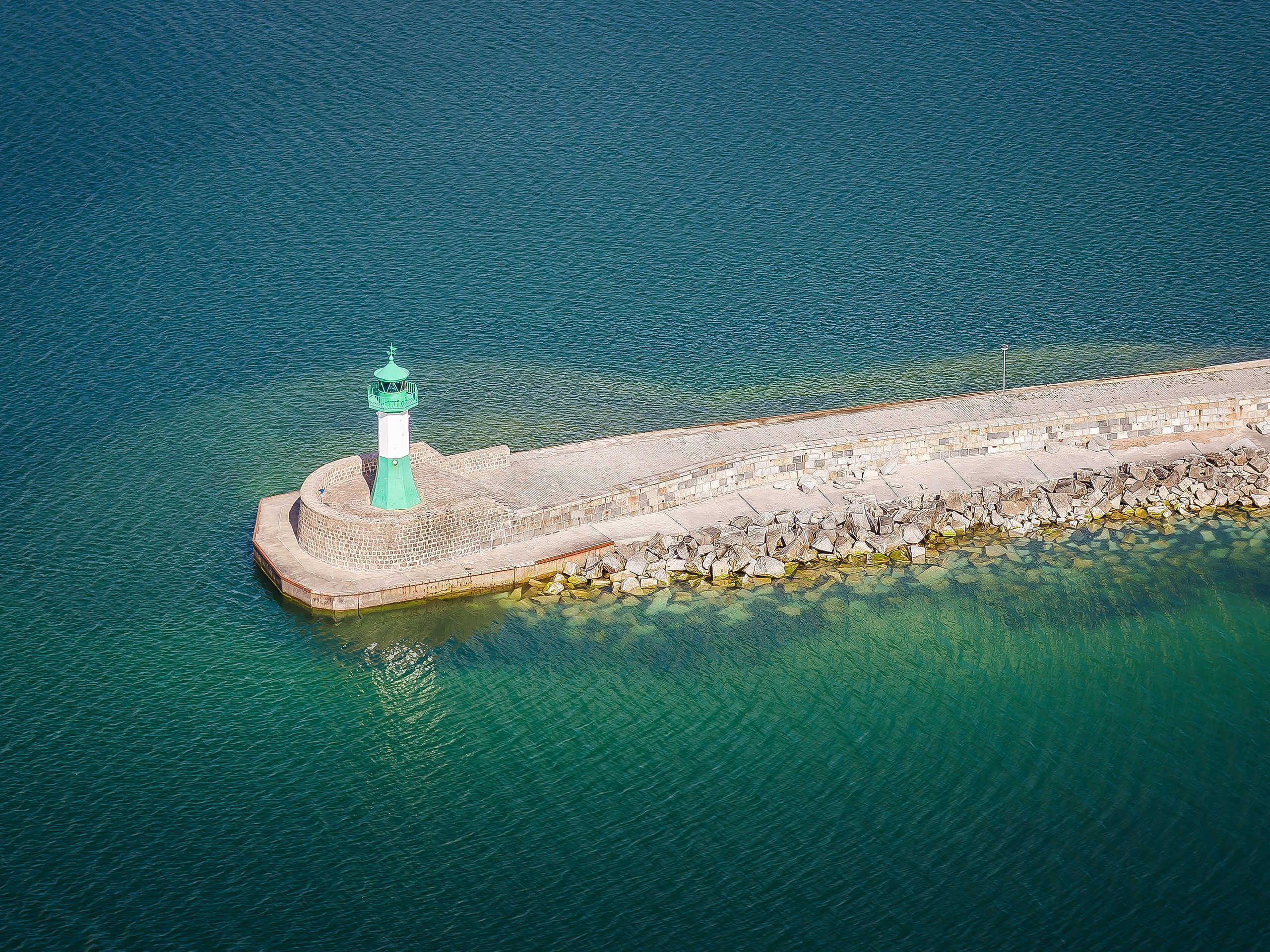 An aerial view of a lighthouse on a rocky breakwater extending into a calm sea.