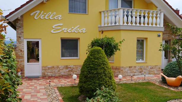 Een gele villa, genaamd 'Villa Erika', met een stenen gevel, een klein balkon en een verzorgde tuin.