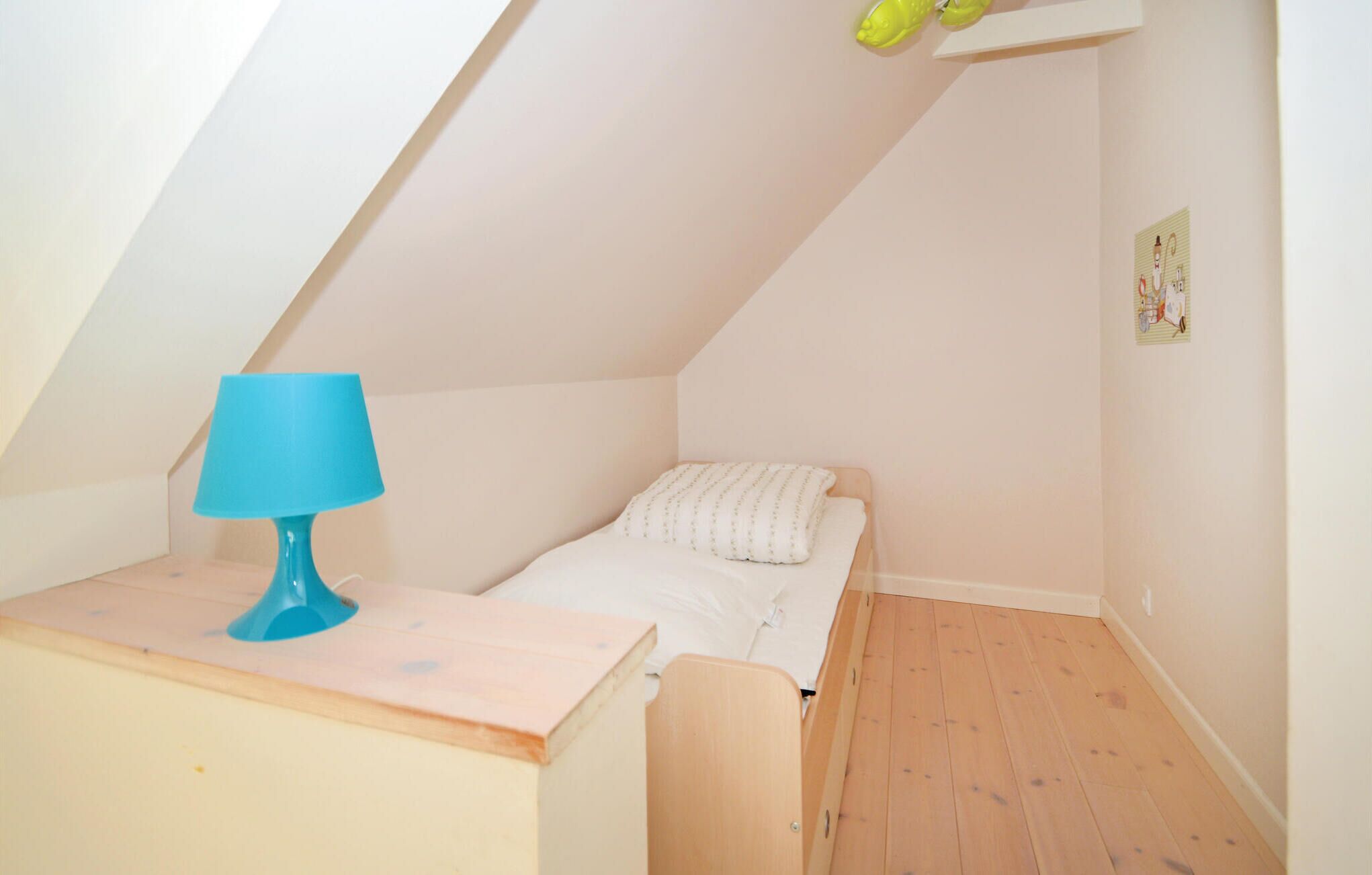 Habitación pequeña con techo inclinado, una cama, una mesita de noche con lámpara y un estante con dos objetos verdes.
