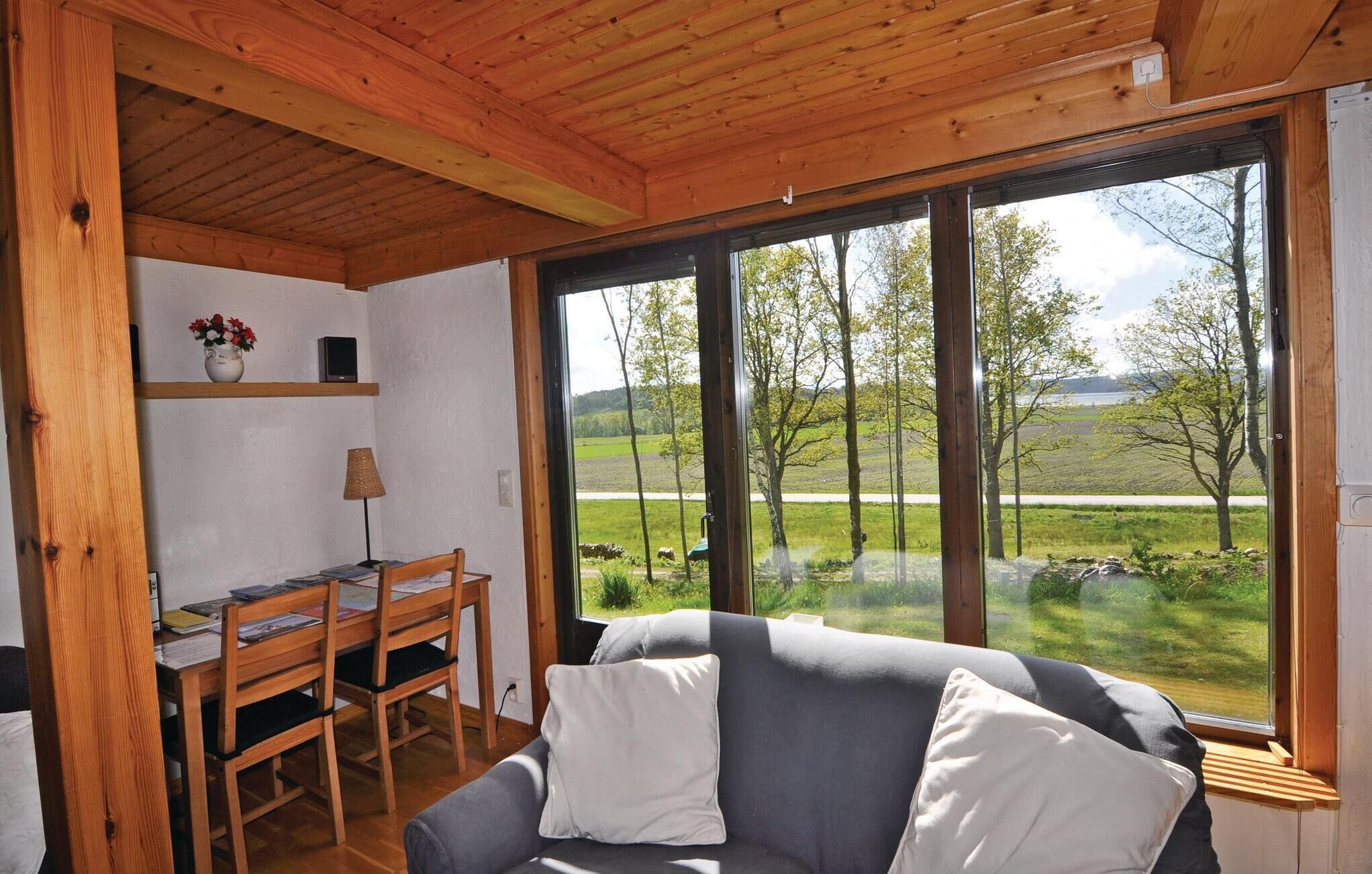 Een knus interieur met een houten plafond, een grijze bank met witte kussens, een houten eettafel met stoelen, en grote ramen met uitzicht op een groen landschap.