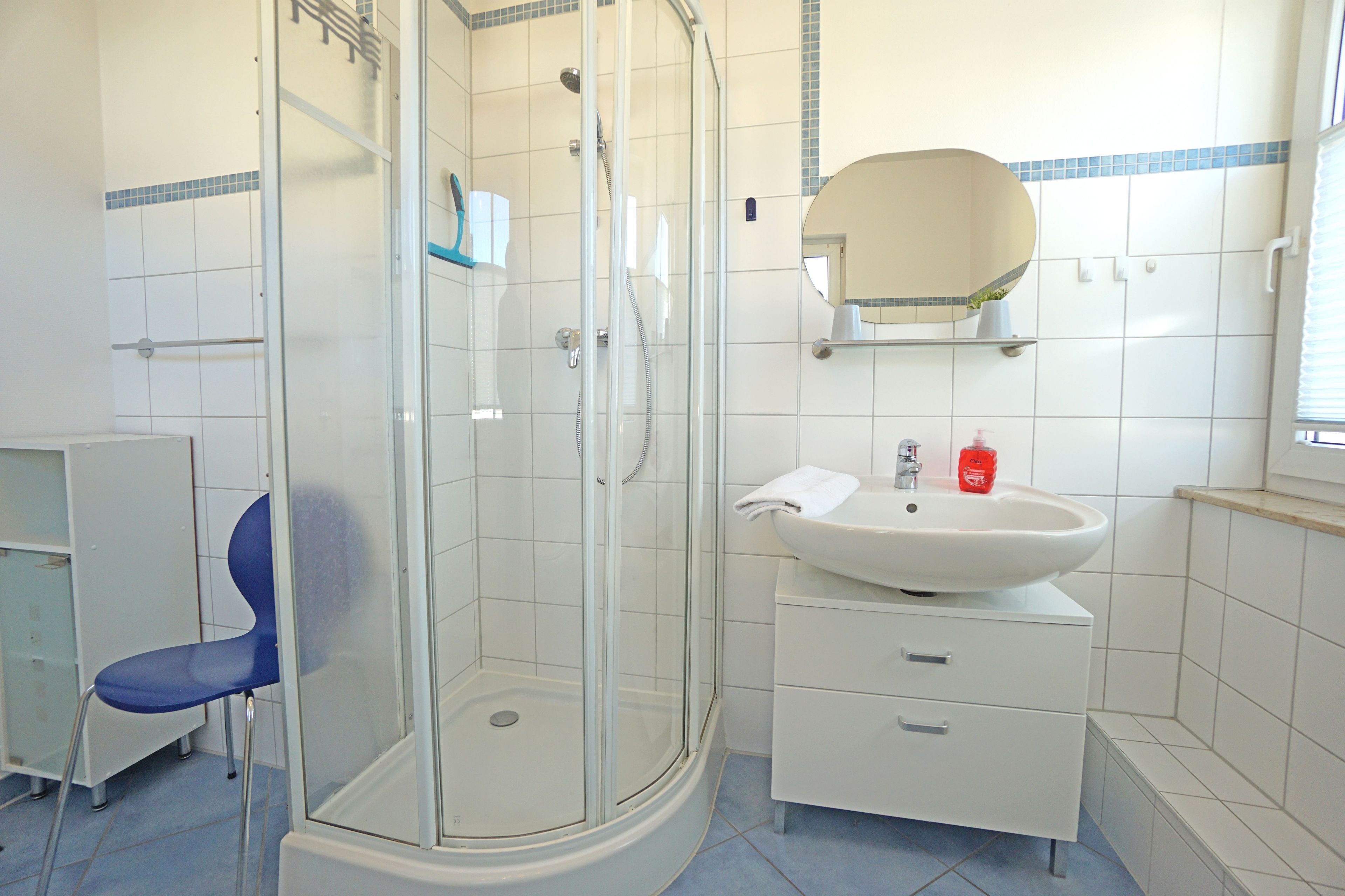 Ein Badezimmer mit einer gläsernen Duschkabine, einem Waschbecken mit Spiegel darüber und einem blauen Stuhl.