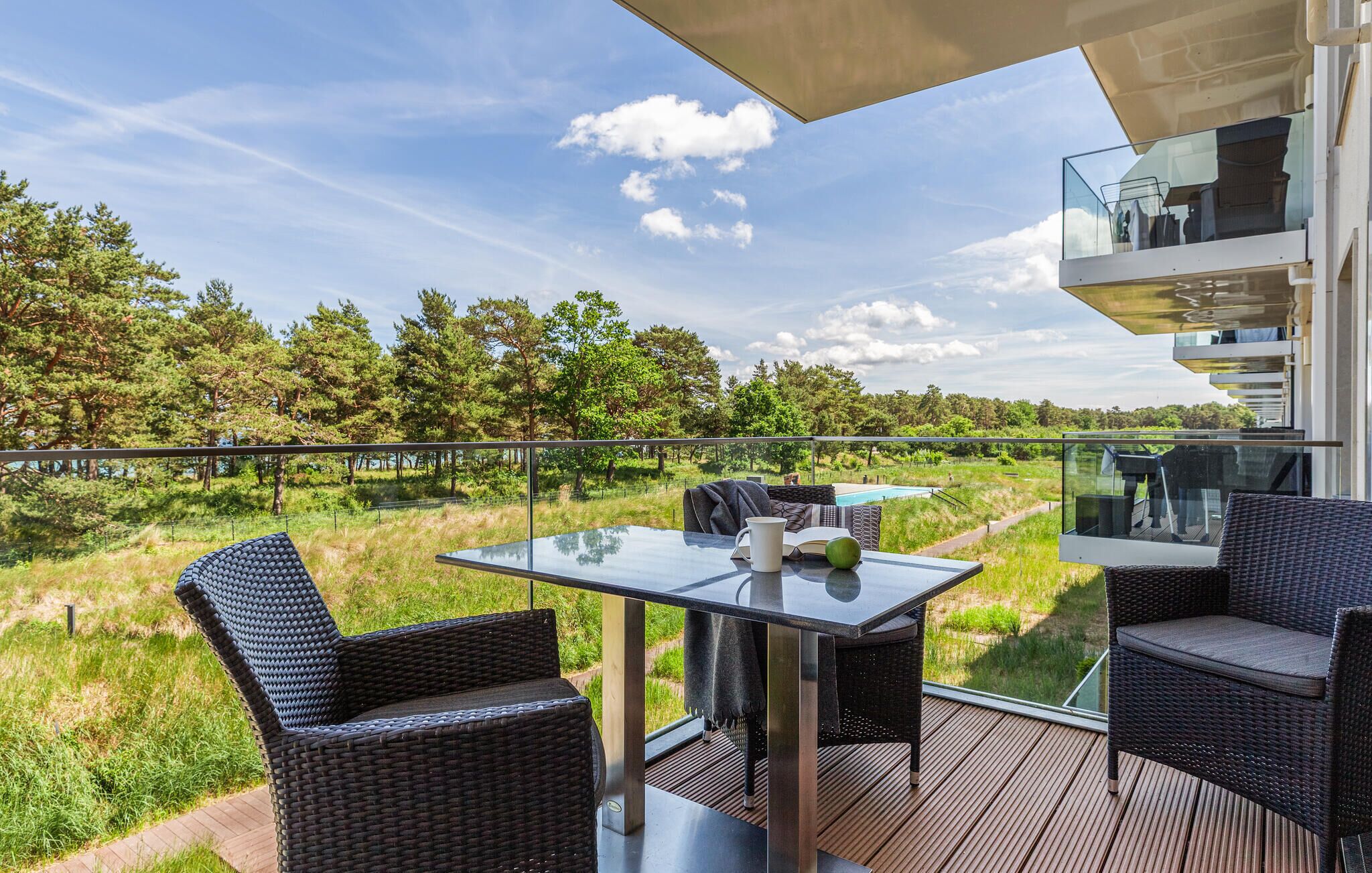 An outdoor balcony with wicker furniture, a glass table, and a view of a natural landscape.