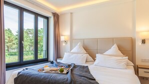 A hotel room with a large window, a bed with white linens, a bedside table, and a tray with breakfast items.