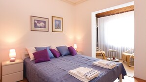 Ein ordentlich bezogenes Bett mit blauer Bettdecke und mehreren Kissen, zwei gerahmte Kunstwerke an der Wand und ein Fenster mit durchsichtigen Vorhängen.