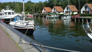 Een jachthaven met aangemeerde boten, een houten steiger en huizen met rode daken op de achtergrond.