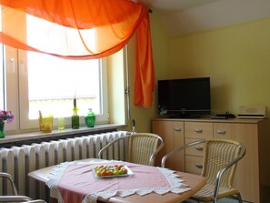 A room with a dining table set for two, a television on a wooden cabinet, and a window with a curtain. - Apartment 35 - Apartments on Usedom (Rankwitz)