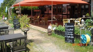 Ein Sitzbereich im Freien mit Tischen und Stühlen unter einem großen roten Sonnenschirm, eine Kreidetafel mit der Speisekarte und ein Gebäude mit einem Strohdach im Hintergrund.