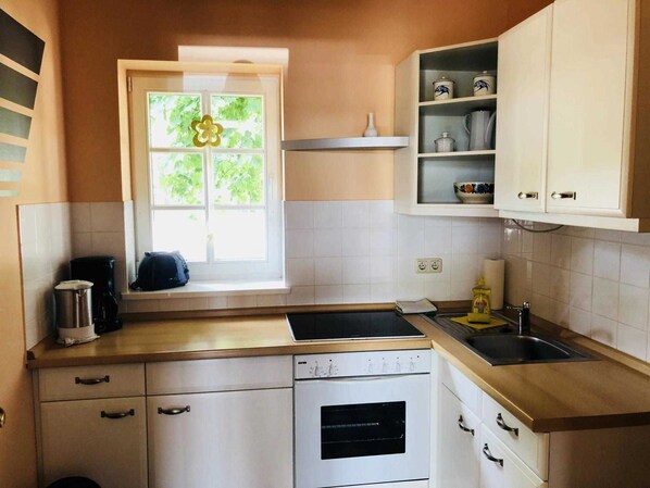A kitchen with white cabinets, a window with a flower decoration, a coffee maker, and a sink. - Apartment 2 - restaurant and boarding Schinkenkrug - Property 50882 (Rostock)