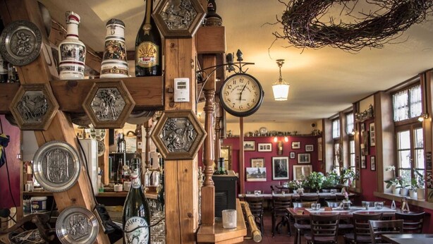 Ein traditionelles Gasthaus mit sechseckigen Holzbögen, die verschiedene Gegenstände präsentieren, einer zentralen Holz-Uhr und einem Essbereich mit Tischen und Stühlen.