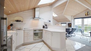 A modern kitchen with white cabinetry, a built-in oven, and a large island. There is a dining area with a table and chairs visible through large windows. - Stunning home in Rønde with kitchen (Rønde)