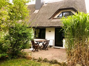 A thatched-roof cottage with a patio table and chairs, surrounded by greenery.