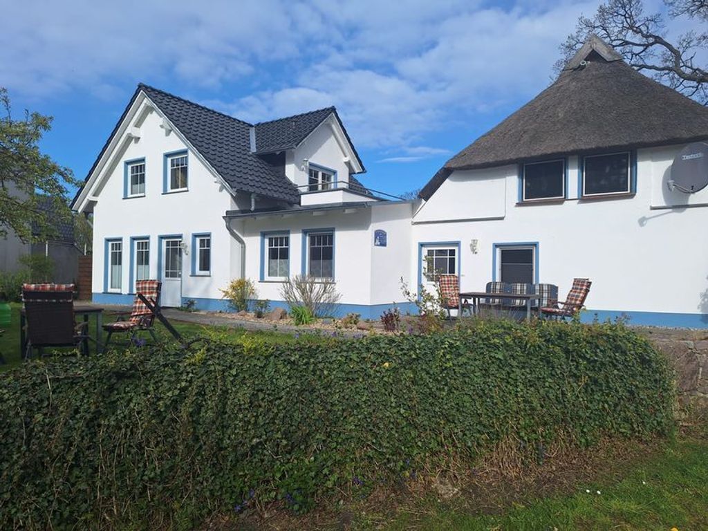 A two-story house with a thatched roof, white exterior, and blue trim. There are outdoor seating areas with chairs and a table.