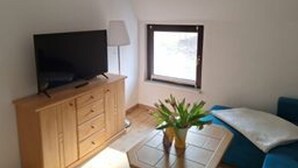 Ein Zimmer mit Fernseher auf einem Holzschrank, einem Fenster mit Blick, einer Pflanze und einem blauen Sofa.