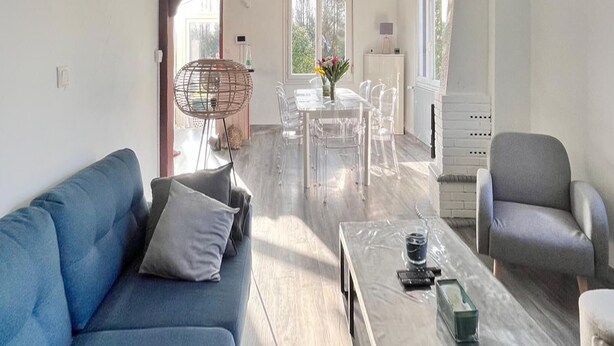 A modern living room with a blue sofa, a grey armchair, a glass coffee table, and a dining area with a clear table and chairs.