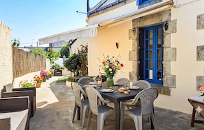 Une terrasse avec une table dressée pour deux personnes, entourée de chaises en osier, d’un vase de fleurs et d’une porte bleue.