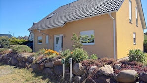 A yellow house with a tiled roof, a door, and a window. There are rocks and plants in front of the house.