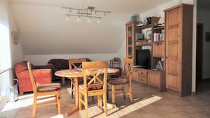 A cozy living room with a wooden dining table, chairs, a sofa, a television, and a bookshelf.