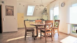 A dining area with a wooden table and chairs, a kitchen in the background, and a window with blinds.