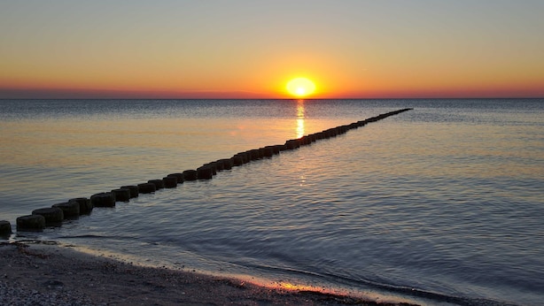 A sunset over a calm sea with a wooden breakwater extending into the water.