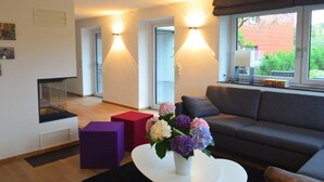 Una sala de estar moderna con un sofá, una mesa de centro con flores y una chimenea. Hay dos taburetes rojo y morado, y una ventana con vistas a follaje.